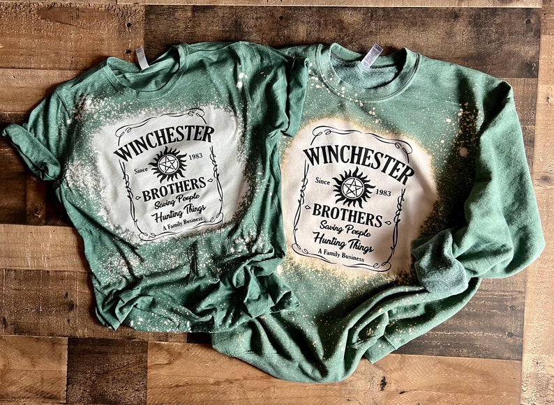 K&ouml;nnte beinhalten: Zwei gr&uuml;ne T-Shirts mit einem Design aus Bleichmittelspritzern. Das Design zeigt ein wei&szlig;es Etikett mit schwarzem Text, der "Winchester Brothers Since 1983 Saving People Hunting Things A Family Business" lautet.