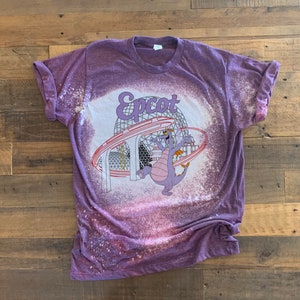 May include: Purple t-shirt with a bleach-splattered design featuring a cartoon dragon and the text "Epcot".