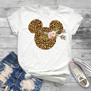May include: White t-shirt with a leopard print Mickey Mouse head design. The Mickey Mouse head is adorned with a floral arrangement and arrows.