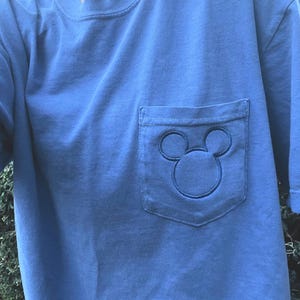 May include: A light blue t-shirt with a pocket. The pocket has an embroidered Mickey Mouse design. The t-shirt has short sleeves and a crew neck.