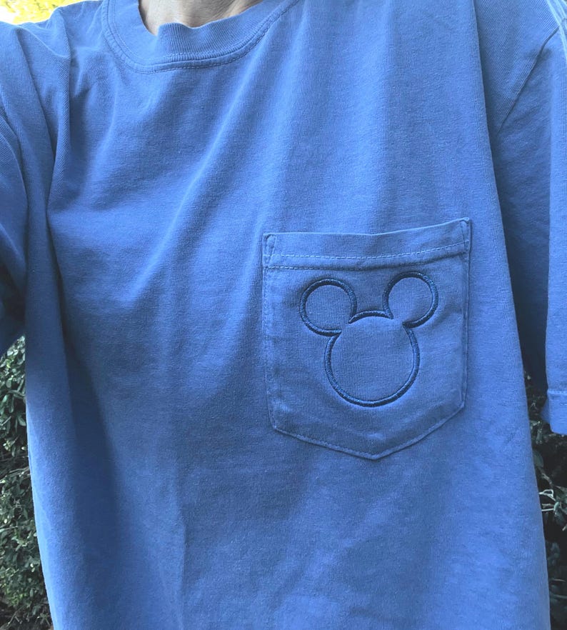 May include: A light blue t-shirt with a pocket. The pocket has an embroidered Mickey Mouse design. The t-shirt has short sleeves and a crew neck.