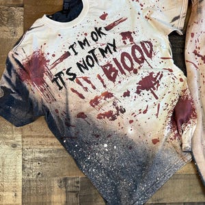 Puede incluir: Una camiseta de manga corta con efecto degradado negro y desteñido, con salpicaduras de pintura roja que parecen sangre. El texto "I'M OK IT'S NOT MY BLOOD" está impreso en negro, junto a un gráfico de una huella de mano roja oscura.