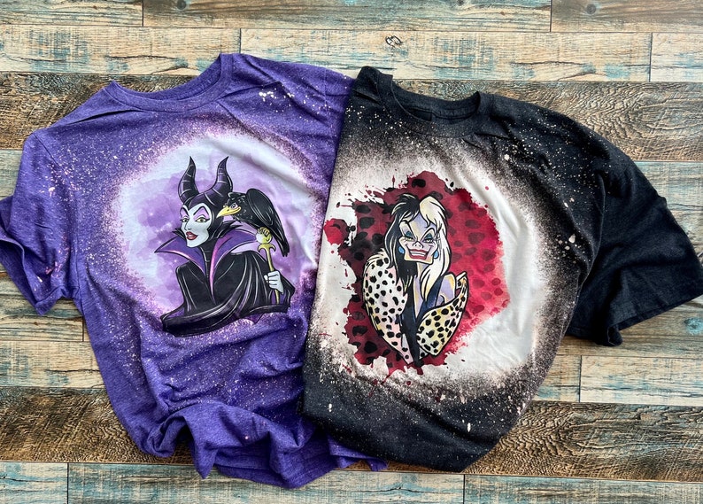 May include: Two bleach-splattered t-shirts featuring cartoon images of Disney villains. The purple shirt has Maleficent with her raven, and the black shirt has Cruella de Vil.