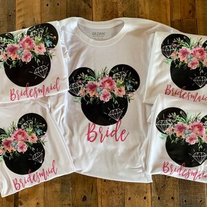 Puede incluir: Cinco camisetas blancas con un diseño de cabeza de Mickey Mouse negro con detalles florales rosas y blancos y formas de diamantes. Las camisetas tienen las palabras "Bride" y "Bridesmaid" impresas.