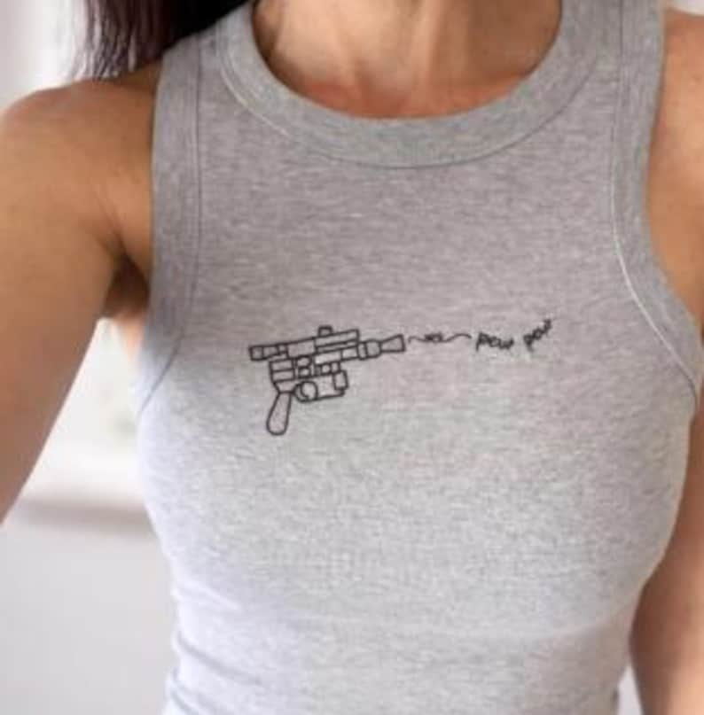 May include: A heather gray sleeveless tank top featuring a black embroidered graphic of a futuristic blaster pistol. The design includes a thin line extending from the barrel with the handwritten text "pew pew" in a cursive script style.