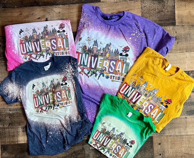 May include: Five bleached t-shirts featuring a graphic print of a castle and the text "Universal Studios". The shirts are in a variety of colours, including pink, purple, yellow, blue, and green.