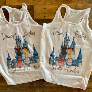 May include: Two white tank tops featuring a graphic print of two girls standing in front of Cinderella's castle. The text "Disney Besties" is printed on the tops, along with the names "Isabelle & Caroline".