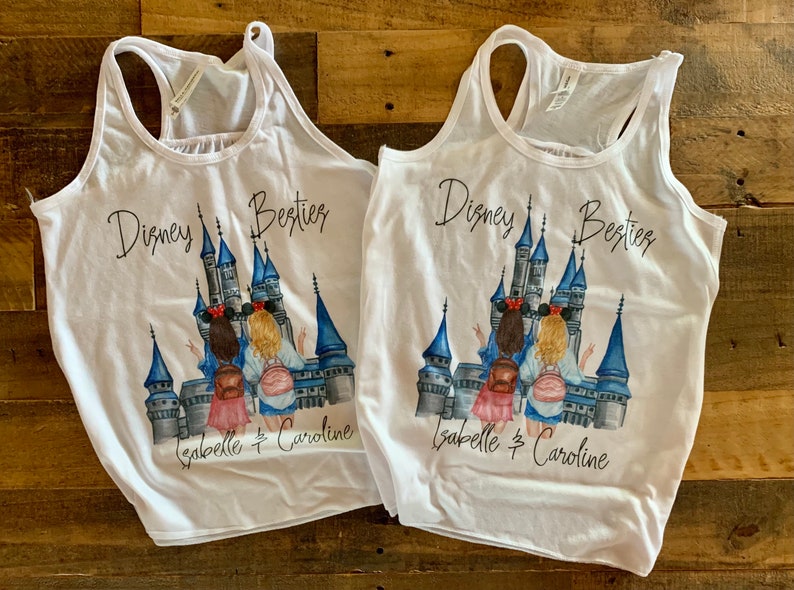 May include: Two white tank tops featuring a graphic print of two girls standing in front of Cinderella's castle. The text "Disney Besties" is printed on the tops, along with the names "Isabelle & Caroline".