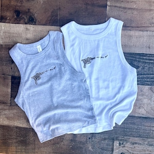 May include: A light gray tank top and a white tank top, both featuring a black embroidered graphic of a blaster pistol with the text "pew pew pew" trailing from the barrel. Each sleeveless shirt has a ribbed crew neckline and a simple cut.