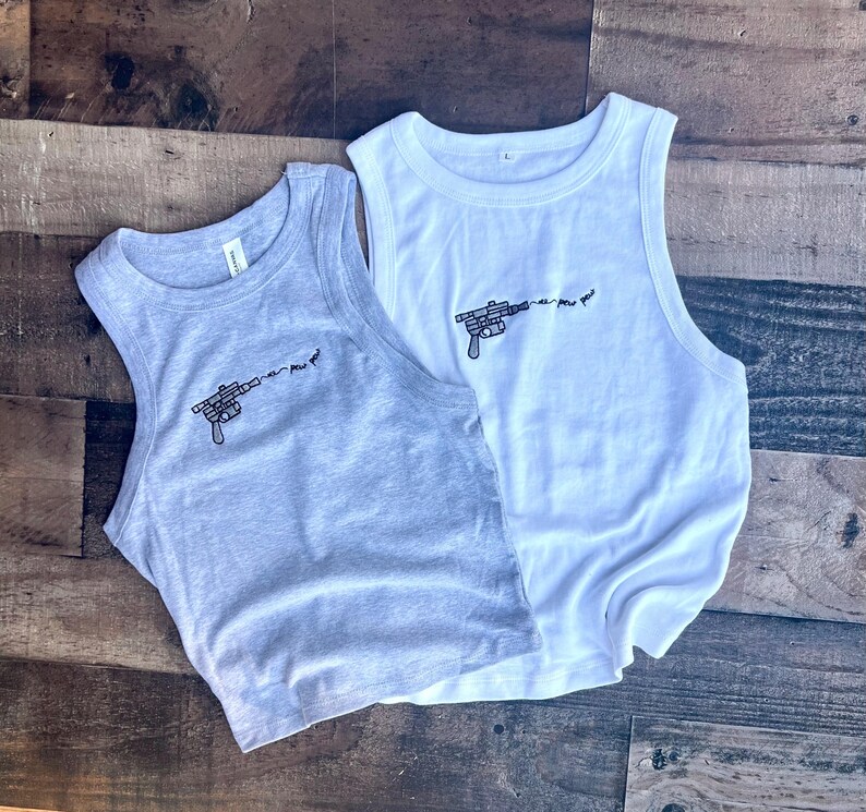 May include: A light gray tank top and a white tank top, both featuring a black embroidered graphic of a blaster pistol with the text "pew pew pew" trailing from the barrel. Each sleeveless shirt has a ribbed crew neckline and a simple cut.