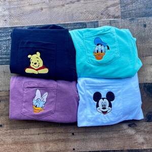 May include: Four t-shirts with pocket detail, in navy, teal, purple, and white. Each has an embroidered Disney character: Winnie the Pooh, Donald Duck, Daisy Duck, and Mickey Mouse. The shirts are folded on a wooden surface.