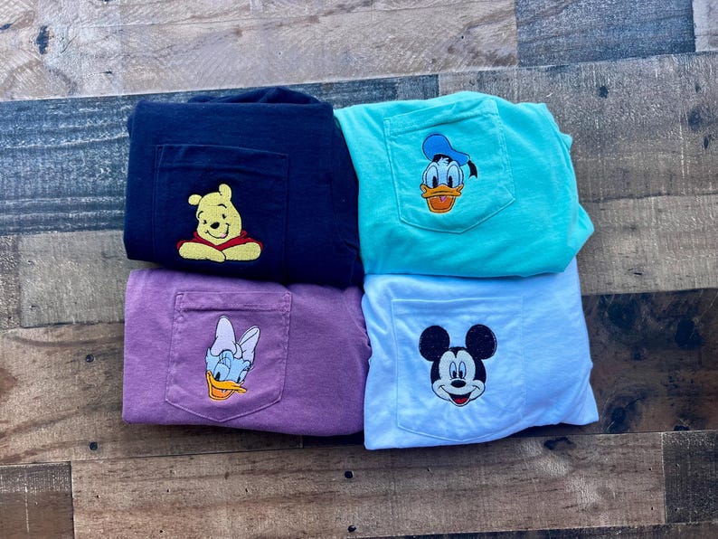 May include: Four t-shirts with pocket detail, in navy, teal, purple, and white. Each has an embroidered Disney character: Winnie the Pooh, Donald Duck, Daisy Duck, and Mickey Mouse. The shirts are folded on a wooden surface.