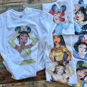 K&ouml;nnte beinhalten: Eine Kollektion wei&szlig;er T-Shirts mit grafischen Illustrationen verschiedener Zeichentrickfiguren, die Mausohr-Haarreifen tragen. Jedes T-Shirt zeigt eine andere Figur mit individueller Haarfarbe, Gesichtsz&uuml;gen und Outfits, darunter gelbe, blaue und goldene Kleider, alles in einem farbenfrohen Cartoon-Stil dargestellt.