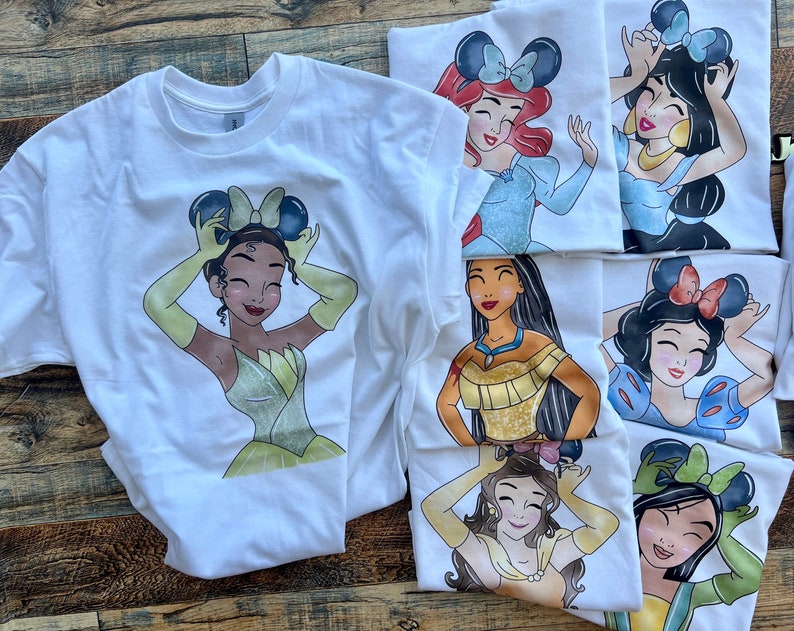 K&ouml;nnte beinhalten: Eine Kollektion wei&szlig;er T-Shirts mit grafischen Illustrationen verschiedener Zeichentrickfiguren, die Mausohr-Haarreifen tragen. Jedes T-Shirt zeigt eine andere Figur mit individueller Haarfarbe, Gesichtsz&uuml;gen und Outfits, darunter gelbe, blaue und goldene Kleider, alles in einem farbenfrohen Cartoon-Stil dargestellt.