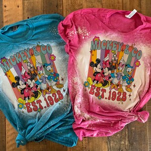 May include: Two bleached t-shirts with a retro-style graphic of Mickey Mouse and friends. The shirts are in blue and pink with the text "Mickey & Co. Est. 1928".