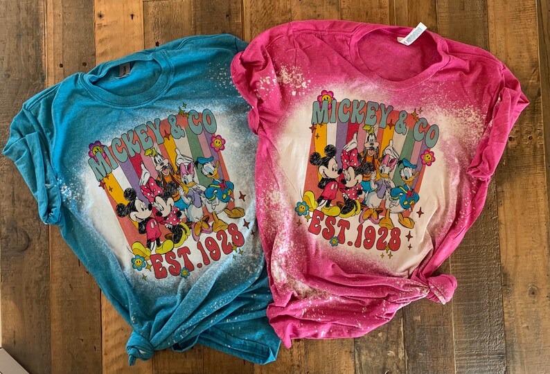 May include: Two bleached t-shirts with a retro-style graphic of Mickey Mouse and friends. The shirts are in blue and pink with the text "Mickey & Co. Est. 1928".