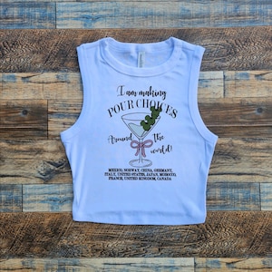 Puede incluir: Top corto blanco con el texto "I am making POUR CHOICES around the world!" y un gr&aacute;fico de copa de martini. La camiseta enumera varios pa&iacute;ses debajo del gr&aacute;fico.