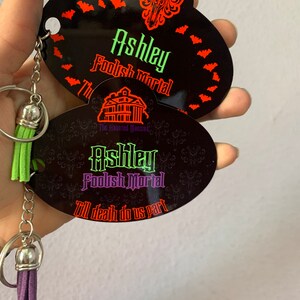 May include: A black and purple Haunted Mansion keychain with a green and purple tassel. The keychain features the text "Ashley Foolish Mortal" and "Till death do us part".