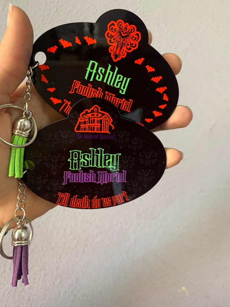 May include: A black and purple Haunted Mansion keychain with a green and purple tassel. The keychain features the text "Ashley Foolish Mortal" and "Till death do us part".