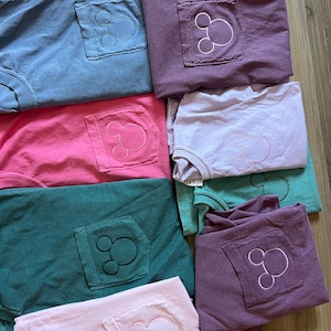 May include: A selection of folded t-shirts in various colours, including blue, pink, green, and purple. Each shirt has a pocket with a Mickey Mouse outline embroidered on it. The shirts are stacked, displaying the embroidered design.