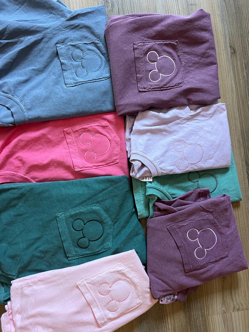 May include: A selection of folded t-shirts in various colours, including blue, pink, green, and purple. Each shirt has a pocket with a Mickey Mouse outline embroidered on it. The shirts are stacked, displaying the embroidered design.