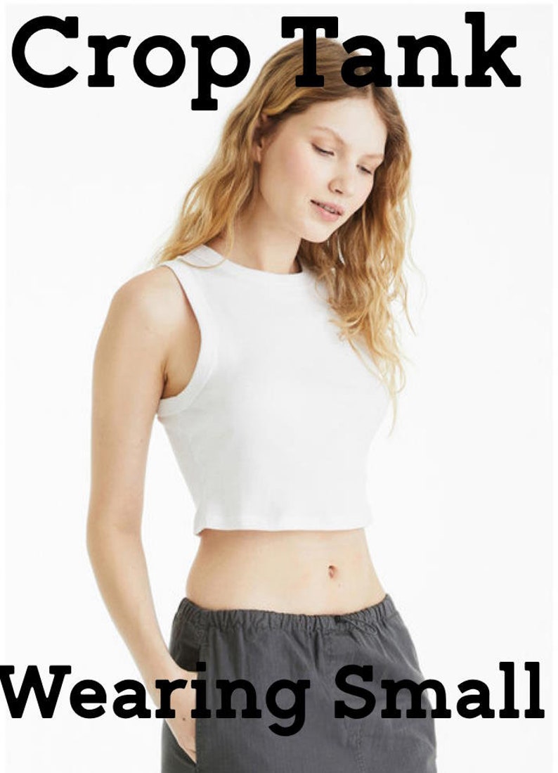 May include: A white crop tank top is shown in this image. The text "Crop Tank" is at the top of the image, and the text "Wearing Small" is at the bottom. The model is wearing gray pants.