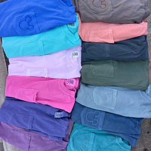 May include: A pile of folded t-shirts in various colours, each with a pocket featuring a stitched Mickey Mouse design. The colours include blue, teal, pink, purple, and grey.