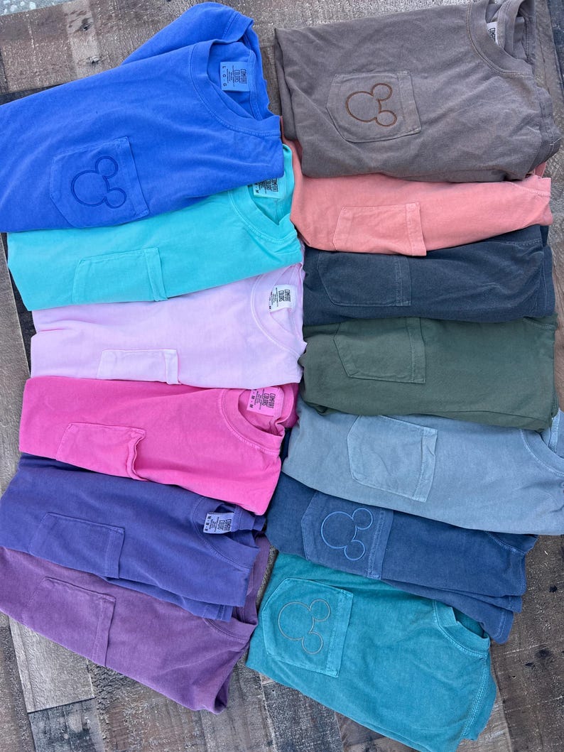 May include: A pile of folded t-shirts in various colours, each with a pocket featuring a stitched Mickey Mouse design. The colours include blue, teal, pink, purple, and grey.