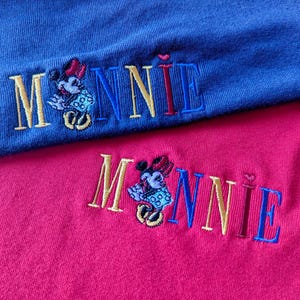 May include: Close-up of two embroidered shirts, one blue and one pink. Each shirt features the name "MINNIE" in gold, blue, and red thread, with an embroidered Minnie Mouse character. The shirts are made of a soft, textured fabric.