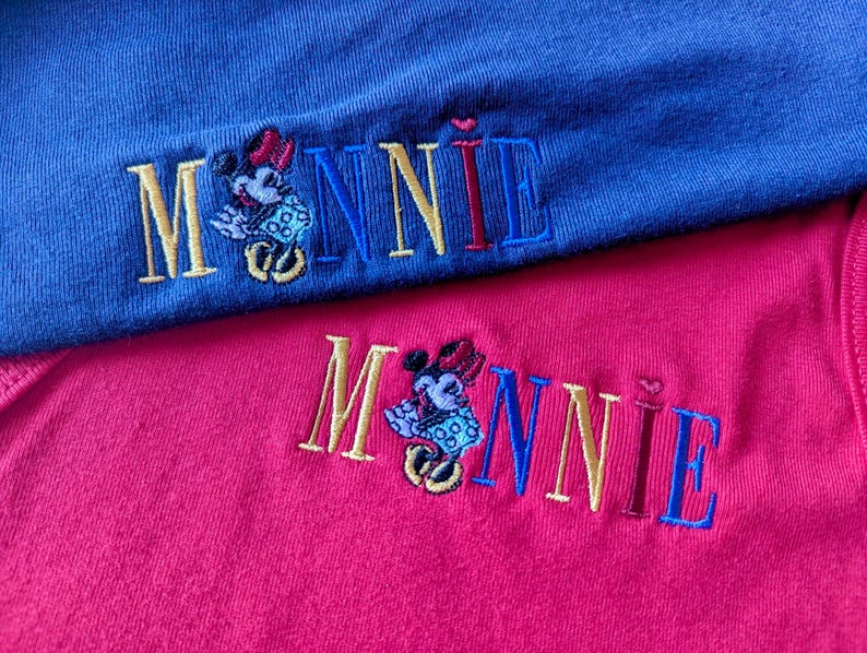 May include: Close-up of two embroidered shirts, one blue and one pink. Each shirt features the name "MINNIE" in gold, blue, and red thread, with an embroidered Minnie Mouse character. The shirts are made of a soft, textured fabric.
