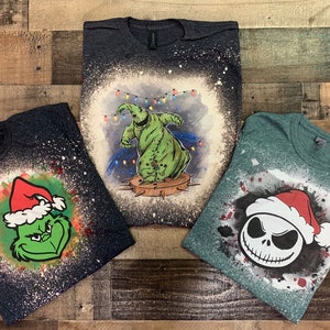 May include: Three t-shirts with bleach-splattered designs featuring Christmas-themed characters. The first shirt is dark blue with a green Grinch wearing a Santa hat. The second shirt is dark gray with a green ghost wearing a Santa hat. The third shirt is green with a black and white Jack Skellington wearing a Santa hat.