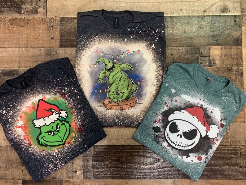 May include: Three t-shirts with bleach-splattered designs featuring Christmas-themed characters. The first shirt is dark blue with a green Grinch wearing a Santa hat. The second shirt is dark gray with a green ghost wearing a Santa hat. The third shirt is green with a black and white Jack Skellington wearing a Santa hat.