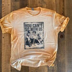 May include: A light yellow t-shirt with a distressed look featuring a black and white image of three girls with the text "You Can't Sit With Us".