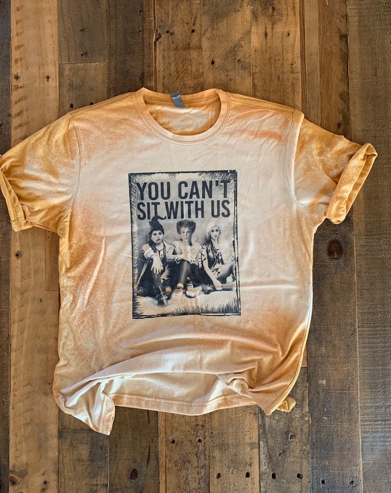 May include: A light yellow t-shirt with a distressed look featuring a black and white image of three girls with the text "You Can't Sit With Us".