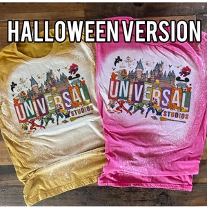 May include: Two bleached t-shirts featuring a Halloween-themed Universal Studios graphic. The shirts are in yellow and pink with a white background. The graphic depicts a castle, Mickey Mouse, and various superheroes.