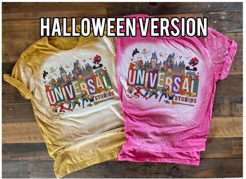May include: Two bleached t-shirts featuring a Halloween-themed Universal Studios graphic. The shirts are in yellow and pink with a white background. The graphic depicts a castle, Mickey Mouse, and various superheroes.