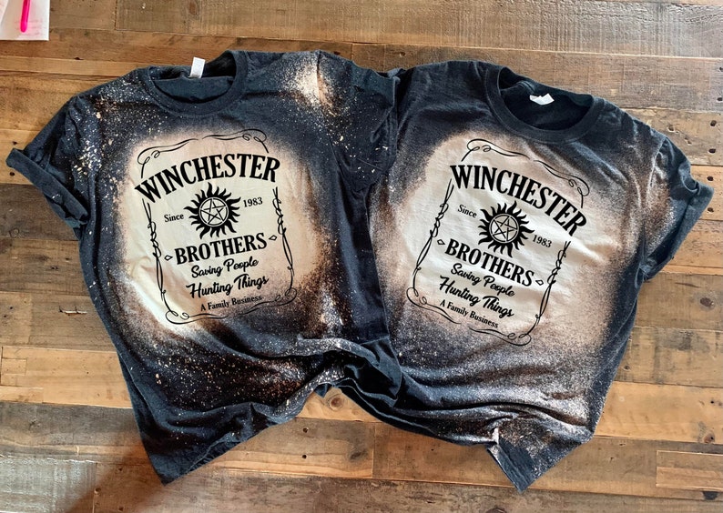 K&ouml;nnte beinhalten: Zwei schwarze T-Shirts mit einem Design aus Bleichmittelspritzern. Die Shirts zeigen ein wei&szlig;es Etikett mit dem Text "Winchester Brothers Since 1983 Saving People Hunting Things A Family Business" und ein Pentagramm-Symbol.