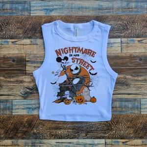 May include: White sleeveless crop top with a graphic print of Jack Skellington from the Nightmare Before Christmas movie. The graphic includes the text "Nightmare on Main Street" and features Jack Skellington holding a ghost with a Mickey Mouse head.