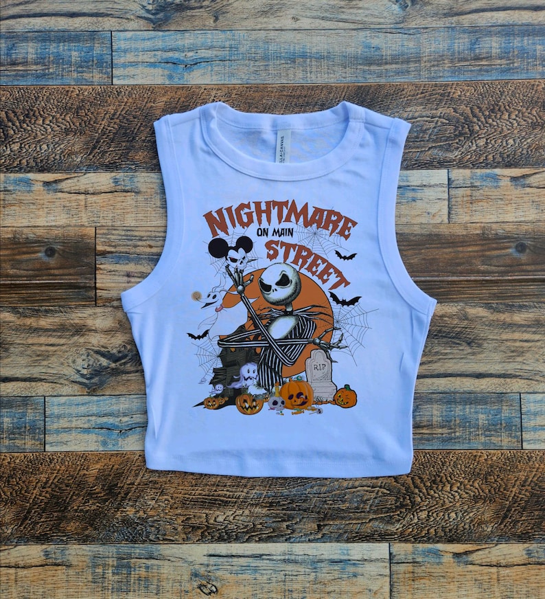 May include: White sleeveless crop top with a graphic print of Jack Skellington from the Nightmare Before Christmas movie. The graphic includes the text "Nightmare on Main Street" and features Jack Skellington holding a ghost with a Mickey Mouse head.