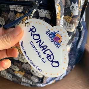 May include: White oval-shaped name tag with the text "RONALDO" in blue, "Victorville, CA" in black, and "WHERE DREAMS COME TRUE" in black. The tag also features a small Disney World logo with a castle and the text "Disneyland" in gold.