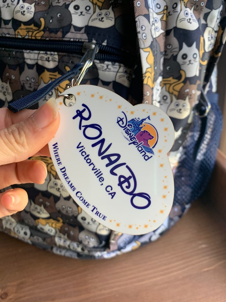May include: White oval-shaped name tag with the text "RONALDO" in blue, "Victorville, CA" in black, and "WHERE DREAMS COME TRUE" in black. The tag also features a small Disney World logo with a castle and the text "Disneyland" in gold.