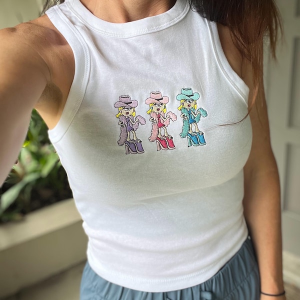 Puede incluir: Una camiseta sin mangas blanca de canalé con un bordado de tres figuras de dibujos animados que llevan sombreros de vaquero, boas y zapatos de plataforma en tonos morado, rosa y azul, situadas horizontalmente en el centro del pecho.