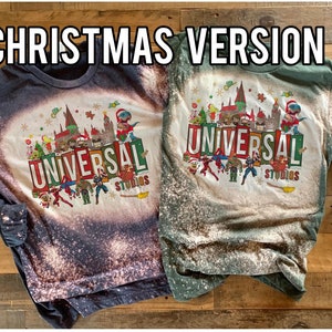 May include: Two long-sleeved shirts with a bleach-splattered design featuring a Christmas-themed Universal Studios logo. The shirts are in dark blue and green.