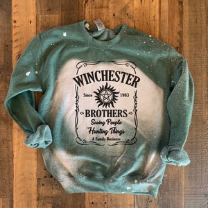 K&ouml;nnte beinhalten: Gr&uuml;nes Sweatshirt mit einem wei&szlig;en Bleichmittel-Design mit dem Text "Winchester Brothers" mit einem Symbol und dem Text "Saving People Hunting Things A Family Business".