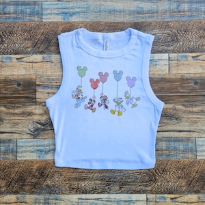 May include: White sleeveless crop top featuring a colorful graphic of cartoon characters holding balloon-shaped balloons. The characters include Mickey Mouse, Donald Duck, and Goofy. The top is made of a soft, lightweight material.