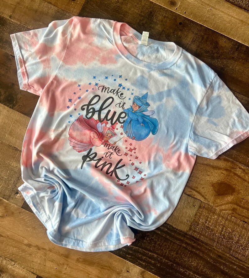 May include: A tie-dye t-shirt with a blue and pink color scheme. The shirt features a graphic of two fairies with the text "Make it blue" and "Make it pink".