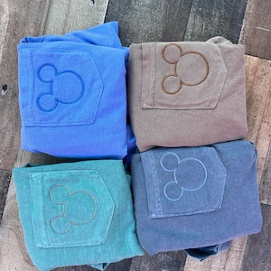 May include: Four pairs of folded trousers in shades of blue, teal, and brown. Each pair has a pocket with an embroidered Mickey Mouse design. The trousers are arranged on a wooden surface.