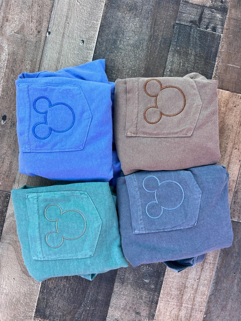 May include: Four pairs of folded trousers in shades of blue, teal, and brown. Each pair has a pocket with an embroidered Mickey Mouse design. The trousers are arranged on a wooden surface.