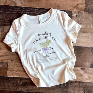 Puede incluir: Camiseta color crema con el texto "I am making POUR CHOICES Around the world!" y un gr&aacute;fico de margarita. La camiseta enumera varios pa&iacute;ses debajo del gr&aacute;fico.