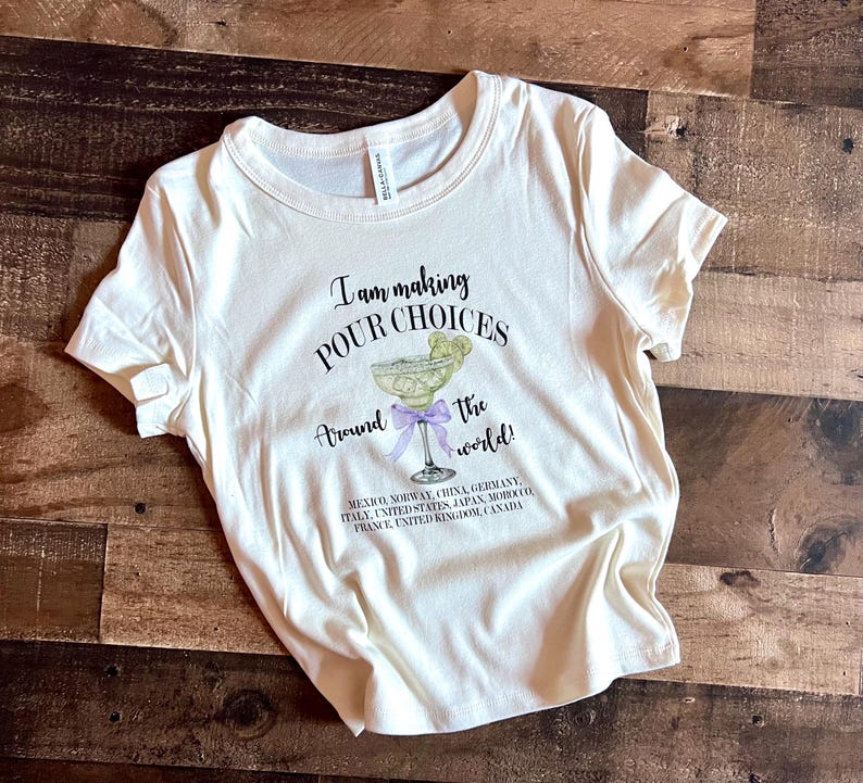 Puede incluir: Camiseta color crema con el texto "I am making POUR CHOICES Around the world!" y un gr&aacute;fico de margarita. La camiseta enumera varios pa&iacute;ses debajo del gr&aacute;fico.